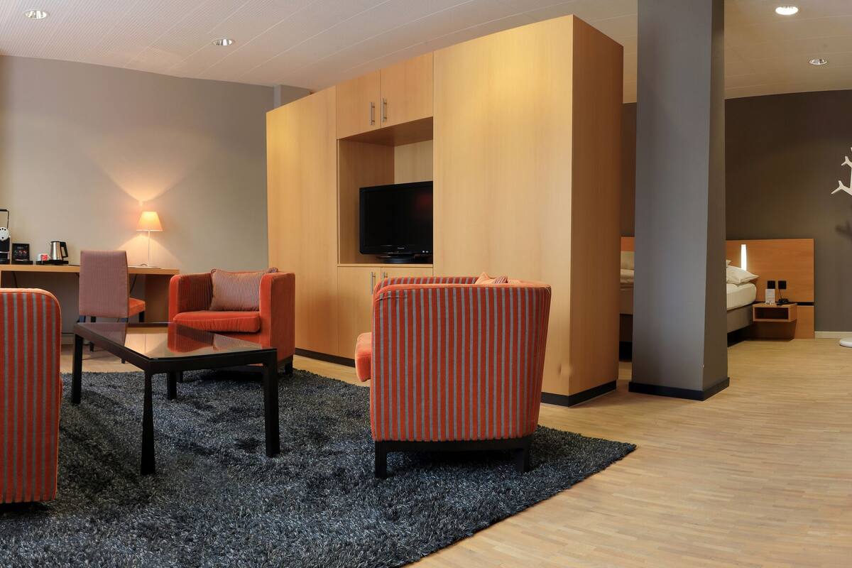 Ein modernes Hotelzimmer mit orangefarbenen Sesseln mit Streifen, einem Glastisch, einem Flachbildfernseher und einer bodengleichen Dusche.