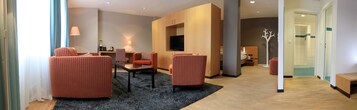 Una habitación de hotel moderna con sillones naranja a rayas, una mesa de centro de vidrio, un televisor de pantalla plana y una cabina de ducha.
