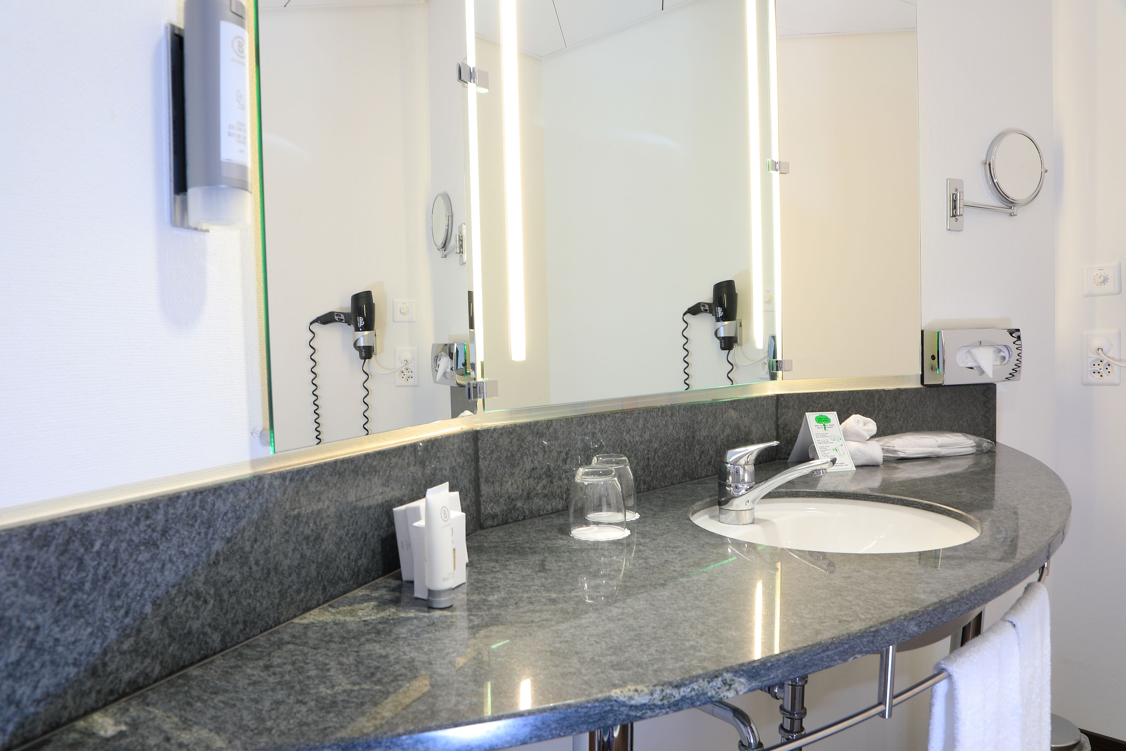 A hotel bathroom with a large mirror, two sinks, a hairdryer, and a towel rack.