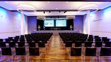 A conference room with rows of chairs, a stage with a presentation screen, and modern lighting fixtures.