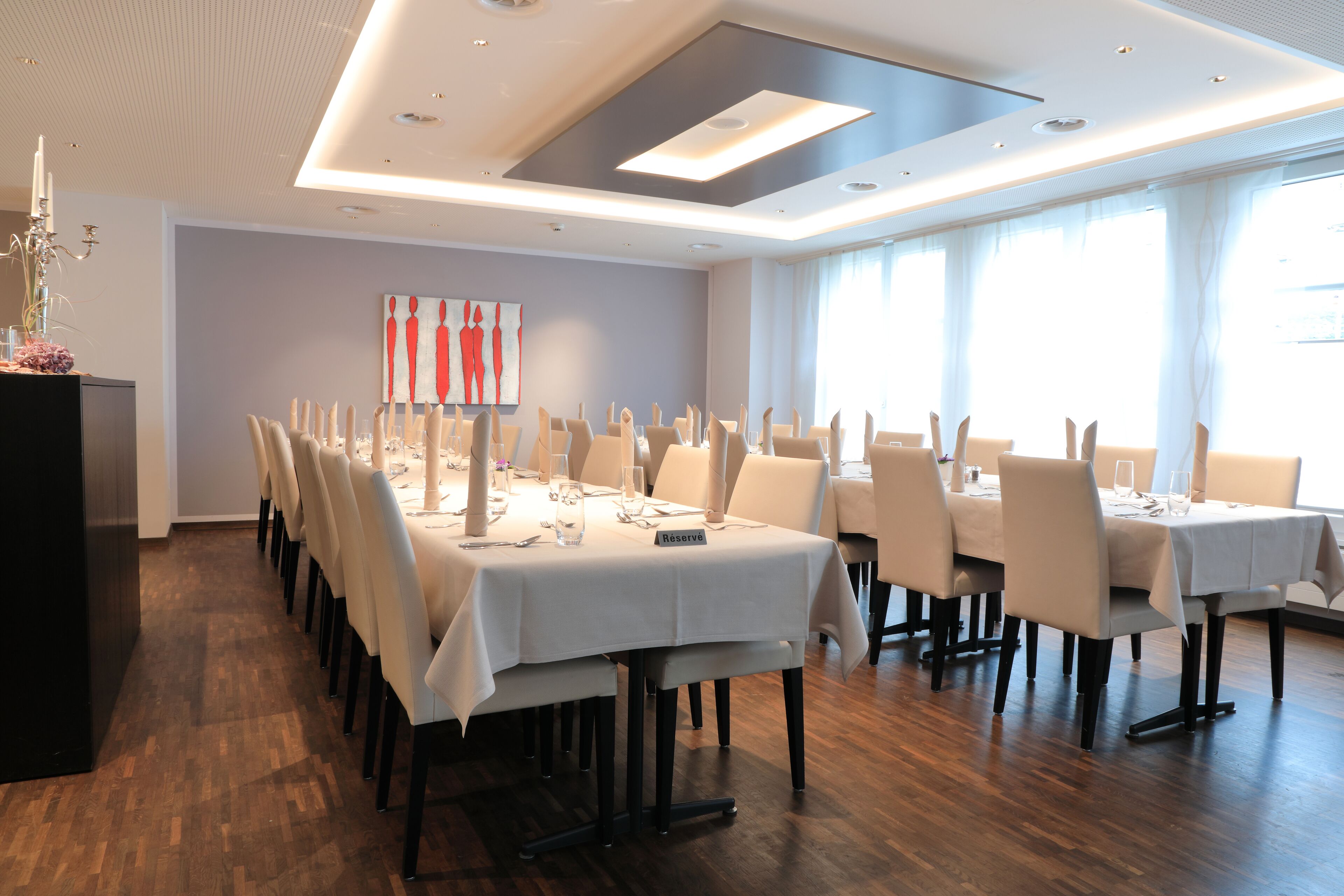 A dining area with multiple tables set for a meal, featuring a modern ceiling design and large windows allowing natural light.