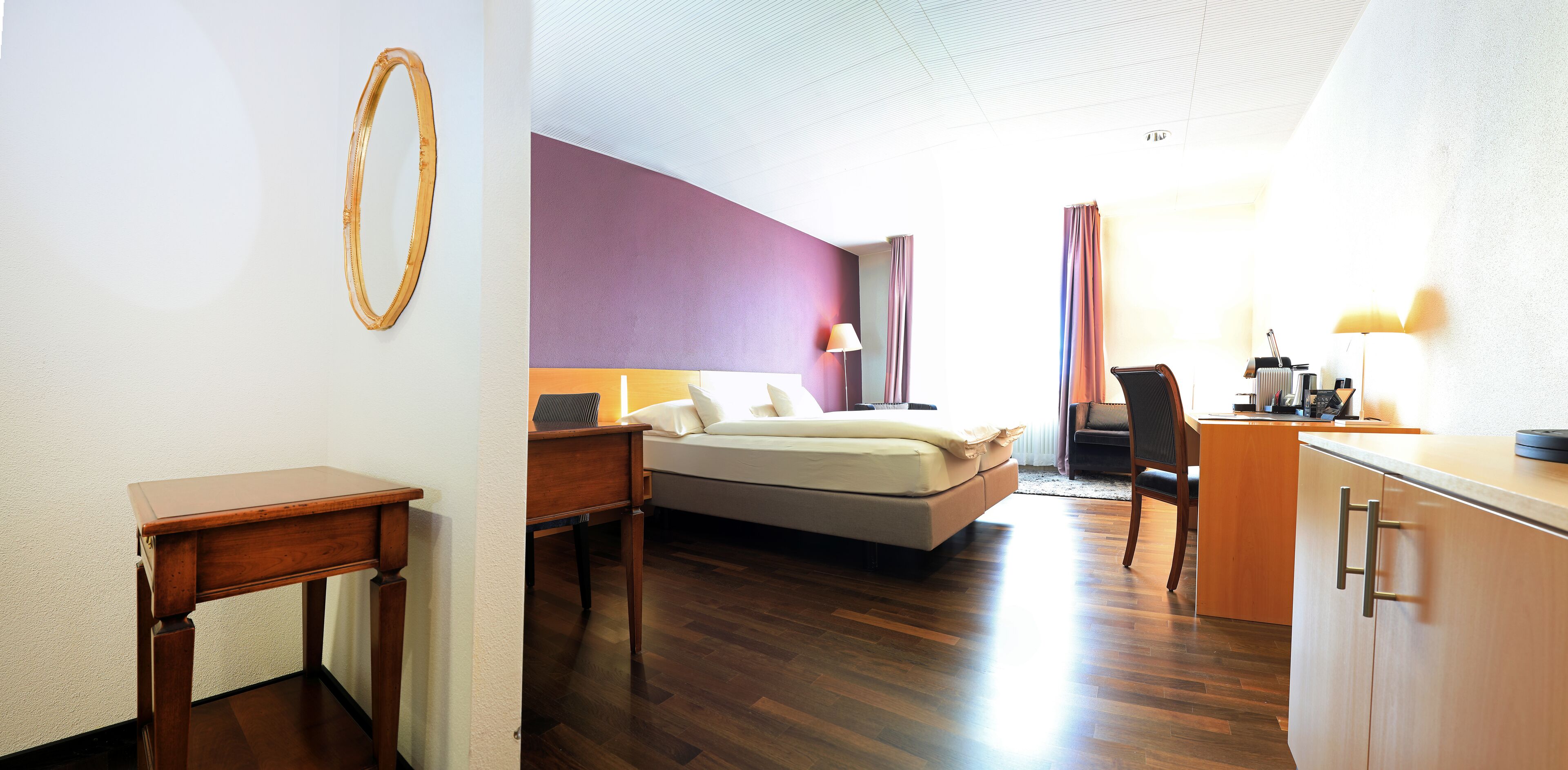 A hotel room with a bed, bedside table, lamp, desk, and a kitchenette area.