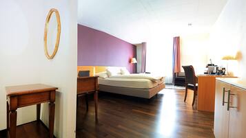 A hotel room with a bed, bedside table, lamp, desk, and a kitchenette area.