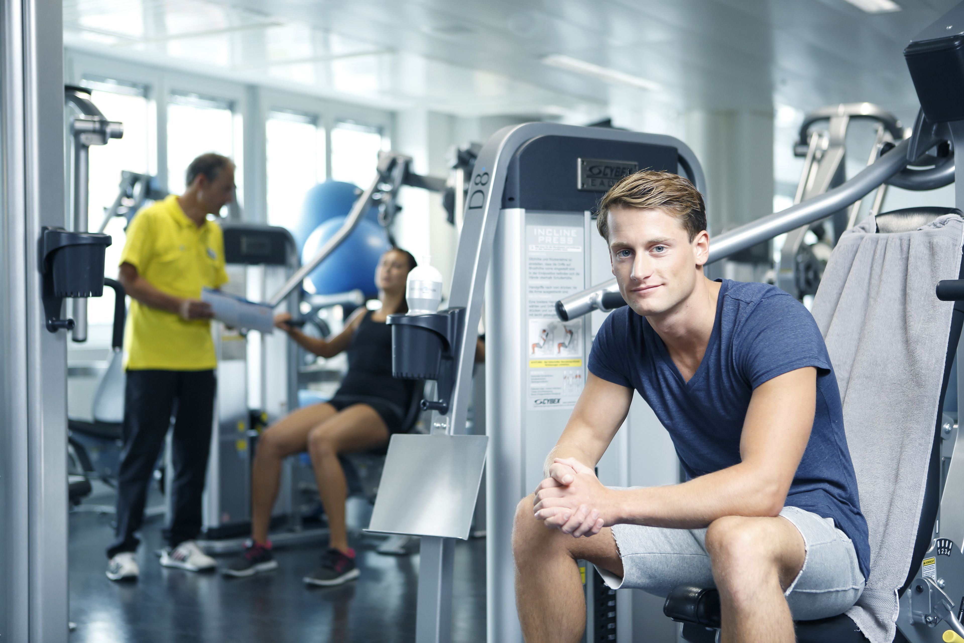 Ein Fitnessstudio mit Trainingsgeräten, ein Mann sitzt an einem Gerät, und ein weiterer Mann steht mit einer Akte herum.
