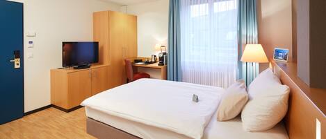 Una habitación de hotel con cama, escritorio, televisión y ventana con cortinas.