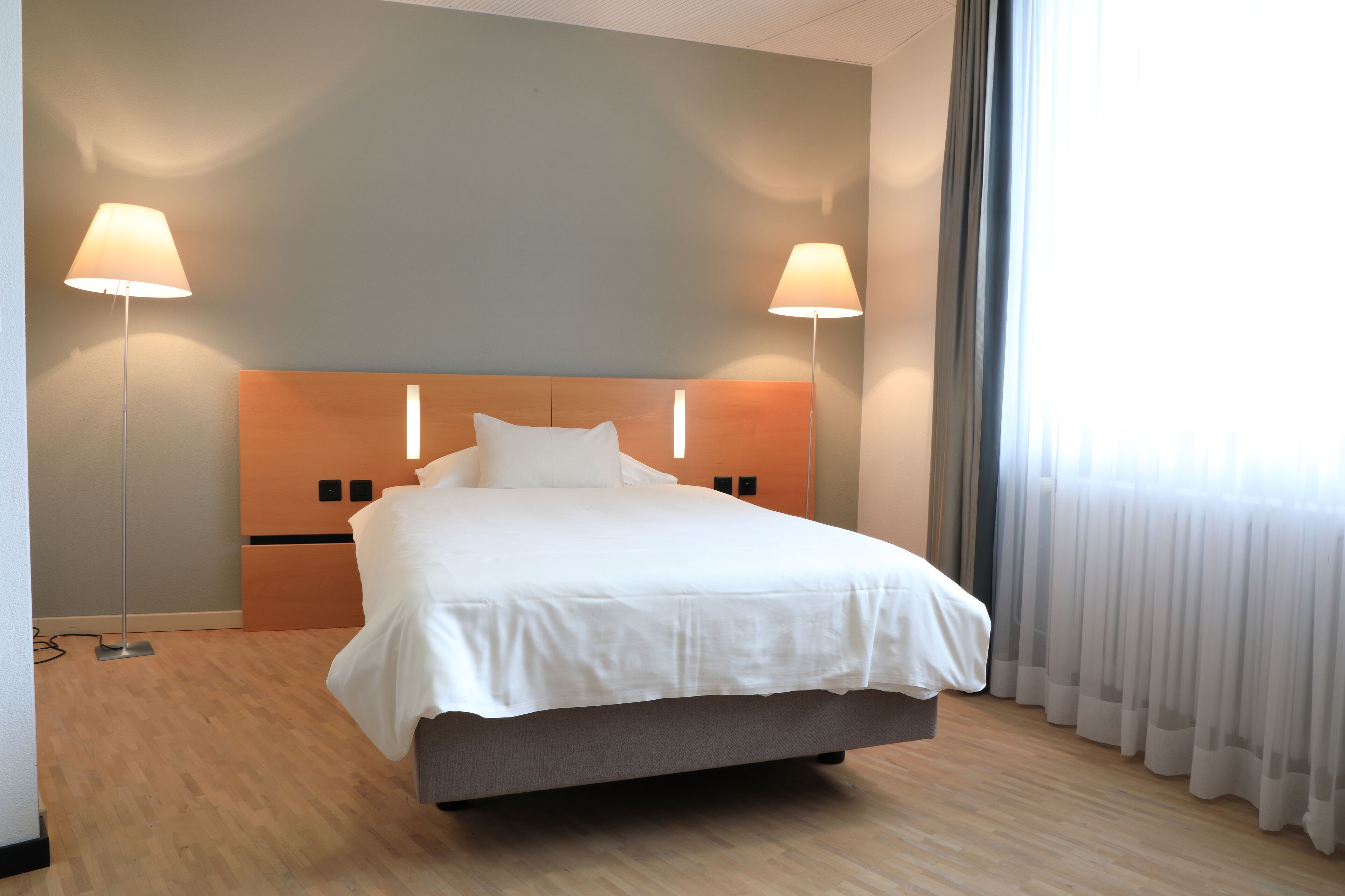 A modern hotel room with a large bed, two bedside lamps, a wooden headboard, and a view of the outdoors through sheer curtains.