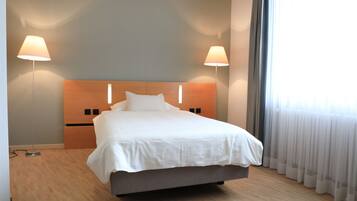 A modern hotel room with a large bed, two bedside lamps, a wooden headboard, and a view of the outdoors through sheer curtains.