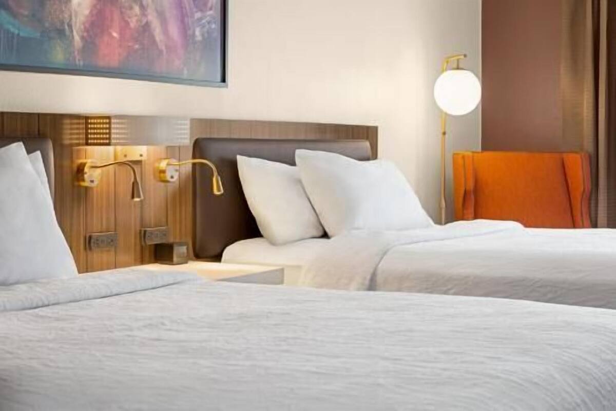Habitación de hotel con dos camas, cabecero de madera, lámpara de pared y un cuadro sobre la cama.