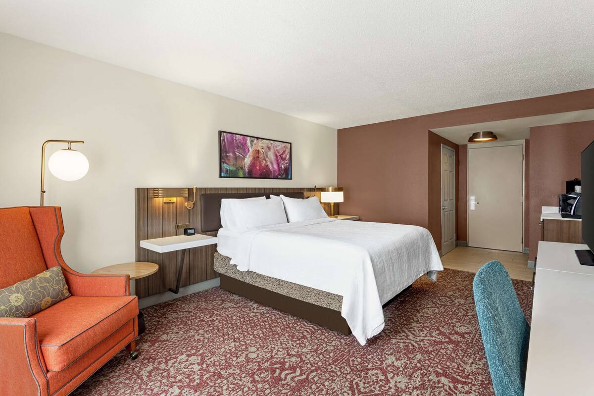 Una habitación de hotel con cama, escritorio, silla, mesita de noche y lámpara de pared.