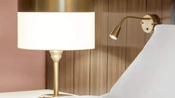 A modern lamp with a white shade and a gold base, positioned on a white bedside table against a wooden wall.