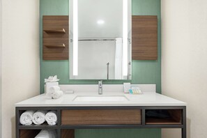 Bathroom features a large mirror above a sink, a vanity with a shelf for towels, and a separate area for bathing.
