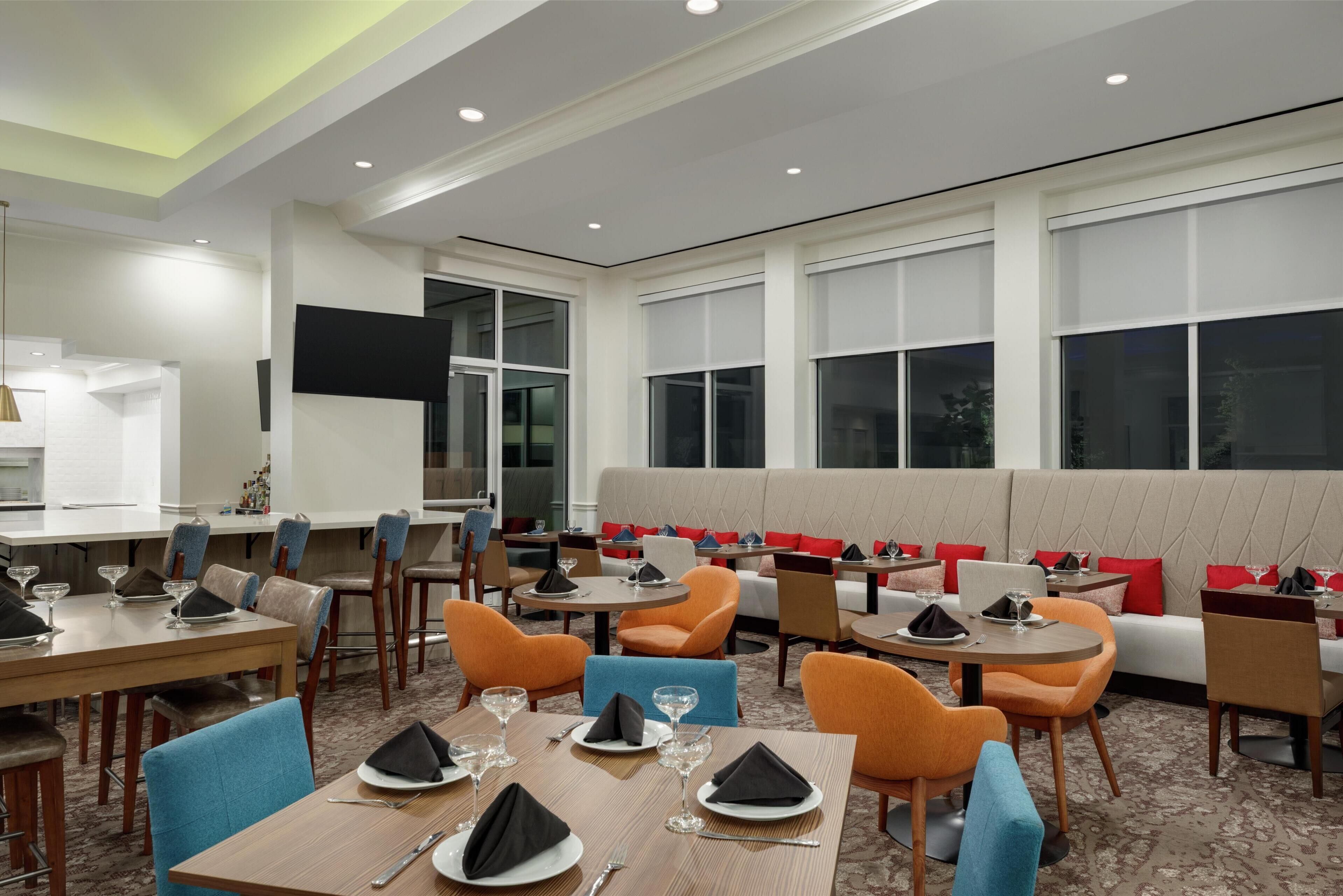 Un comedor de hotel con varias mesas y sillas, una zona de estar con un banco acolchado y amplias ventanas.