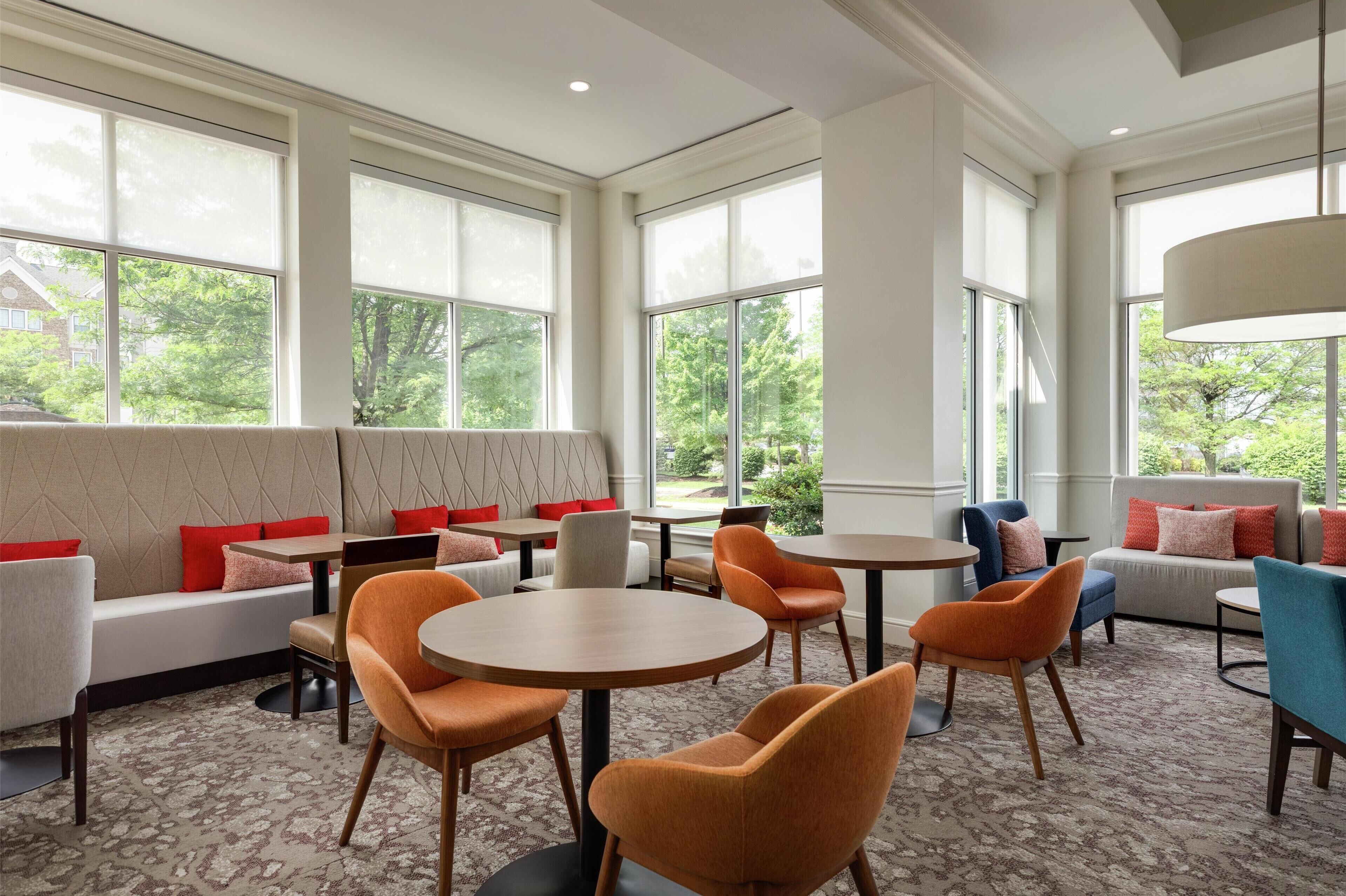 A modern lounge area with seating and tables, large windows with blinds, and a view of greenery outside.