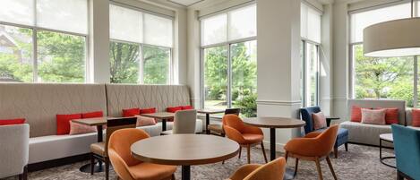 A modern lounge area with seating and tables, large windows with blinds, and a view of greenery outside.