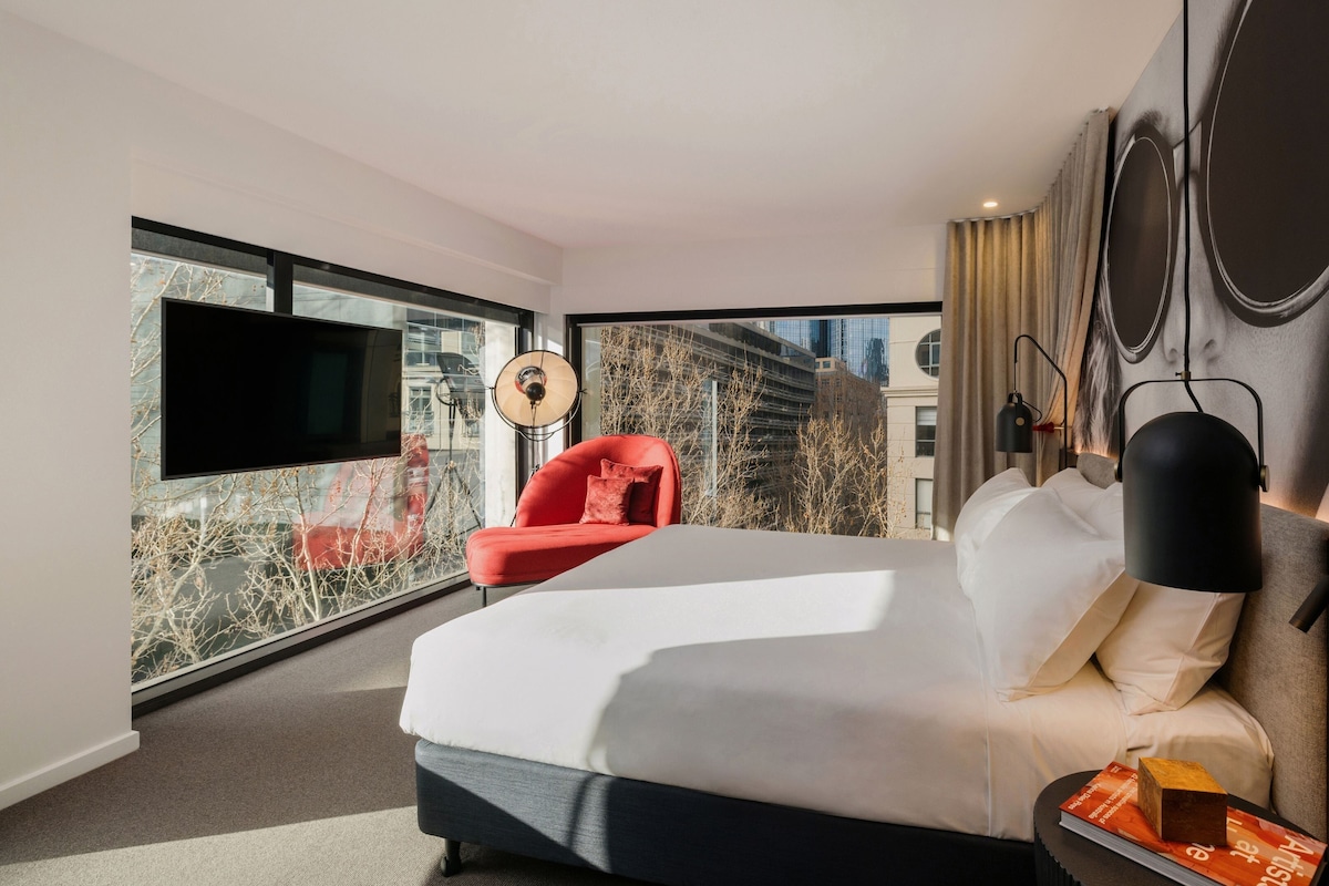 A modern hotel room with a large bed, a red armchair, a flat-screen TV, and a city view through floor-to-ceiling windows.