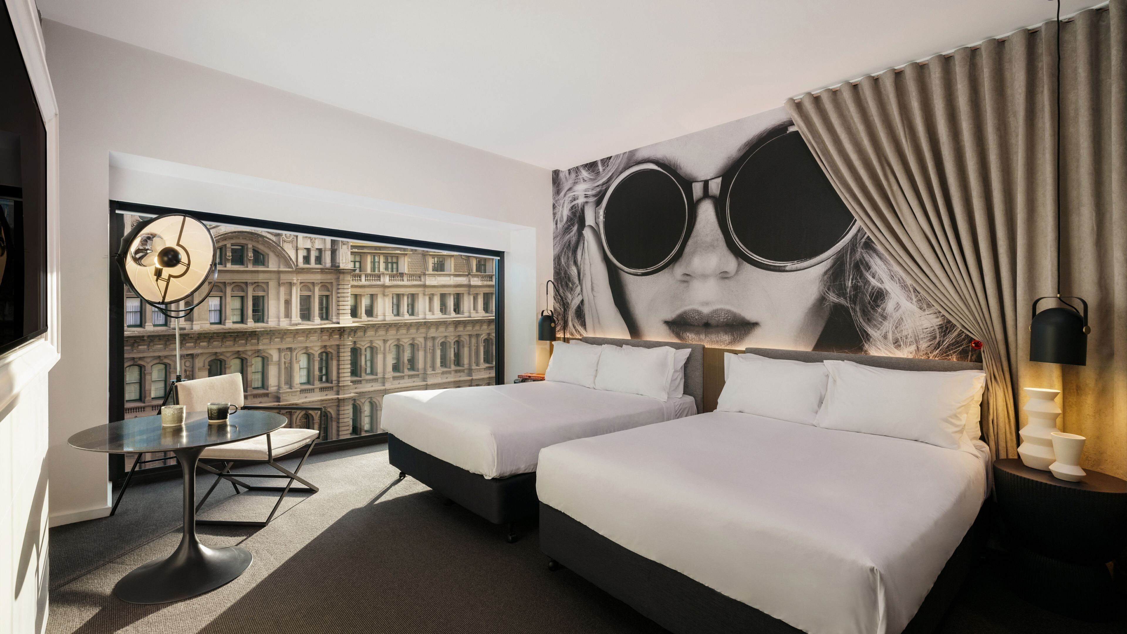 A hotel room with two beds, a desk, and a chair. There is a large window with a view of a historic building.