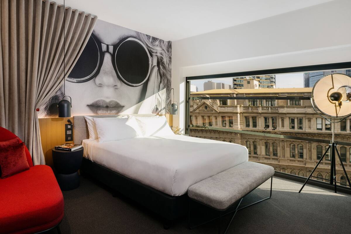 Ein Hotelzimmer mit einem Bett, einem roten Sessel, einem großen Fenster mit Blick auf die Stadt und einem Wandbild.