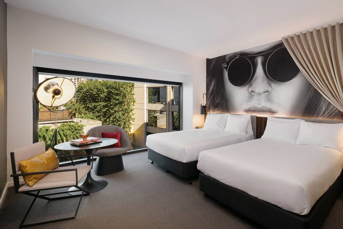 Ein modernes Hotelzimmer mit zwei Betten, einem kleinen Tisch mit Stuhl, einem großen Fenster mit Blick auf Grünflächen und einem einzigartigen Kunstwerk an der Wand.
