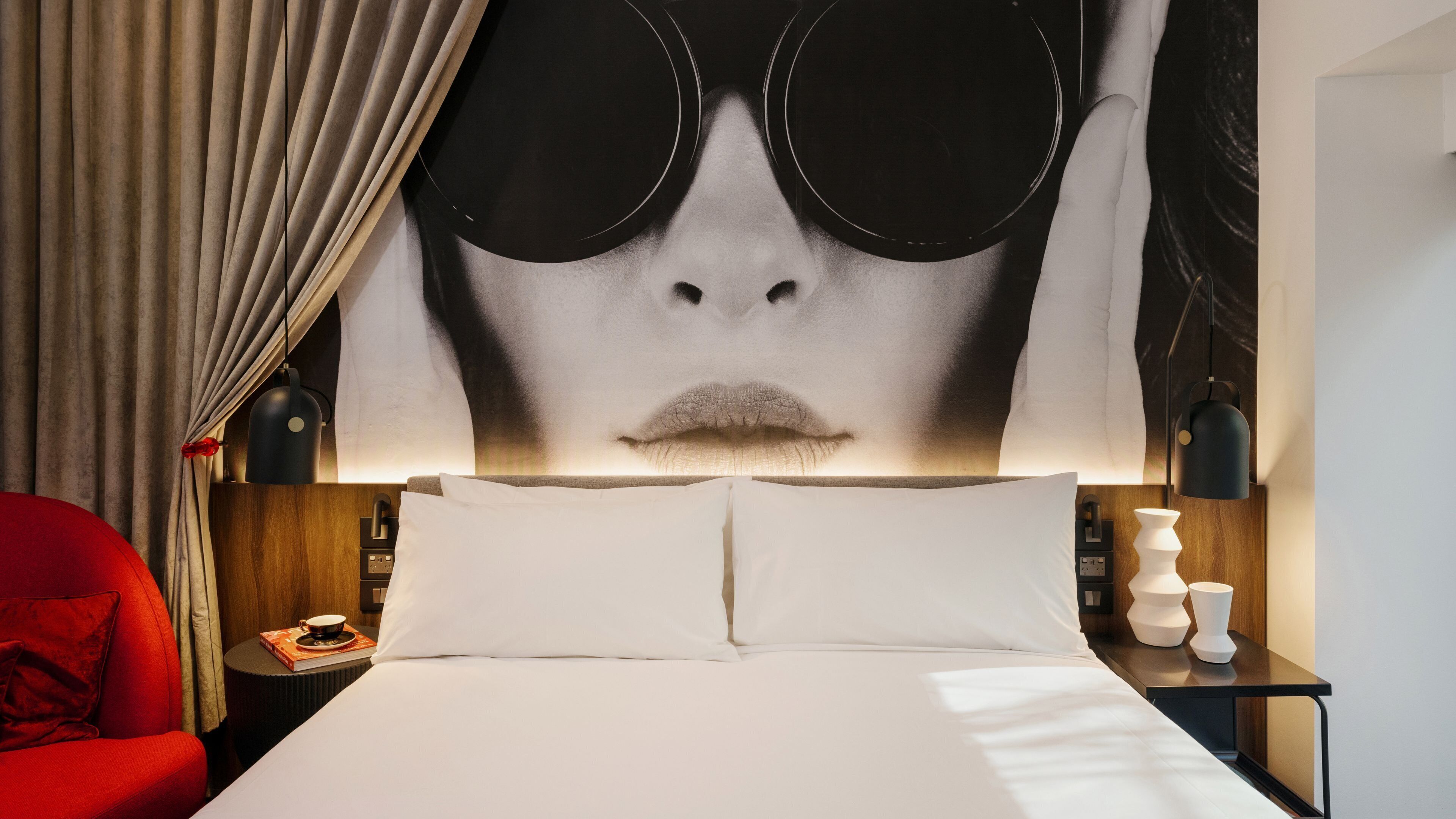 A hotel room with a large bed, a red armchair, a nightstand with a lamp, and a wall with a large artwork.