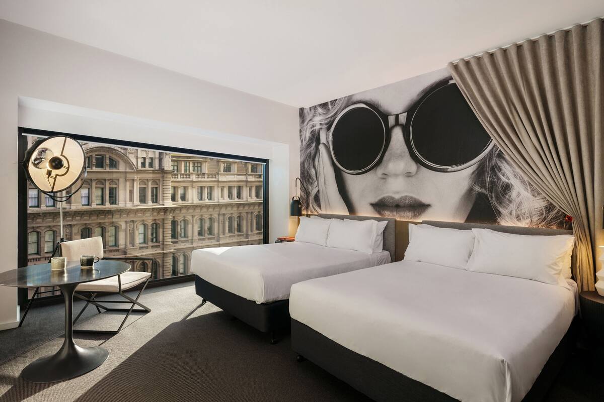 Ein Hotelzimmer mit zwei Betten, einem Schreibtisch und einem Stuhl. Ein großes Fenster bietet Blick auf ein historisches Gebäude.