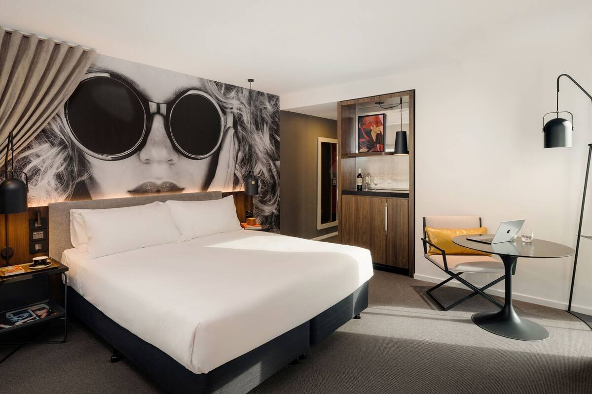 Ein modernes Hotelzimmer mit einem großen Bett, einem Schreibtisch mit einem Laptop, einer Stehlampe und einem an der Wand befestigten Fernseher.