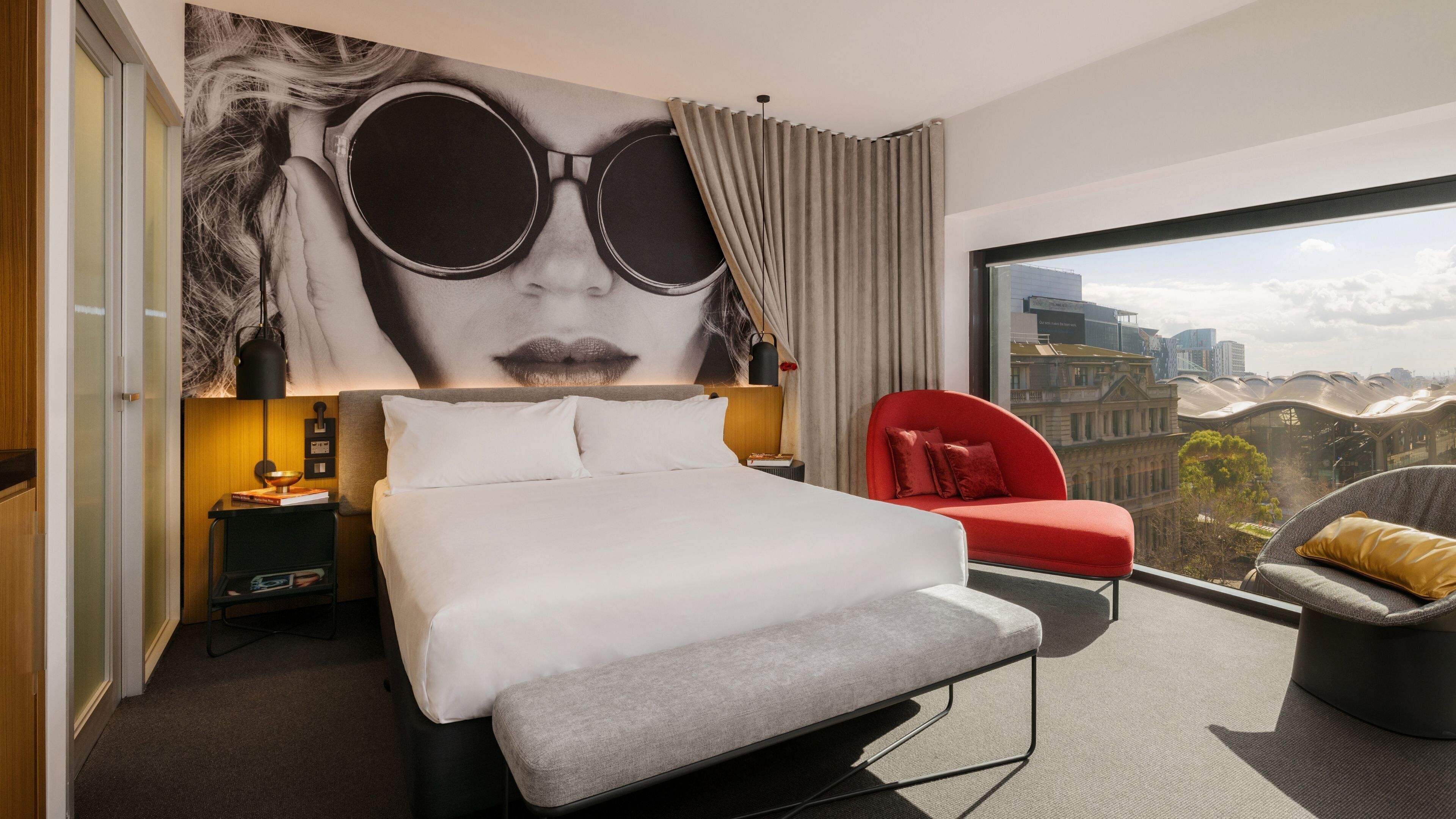 A modern hotel room with a large bed, a red chair, and a city view.