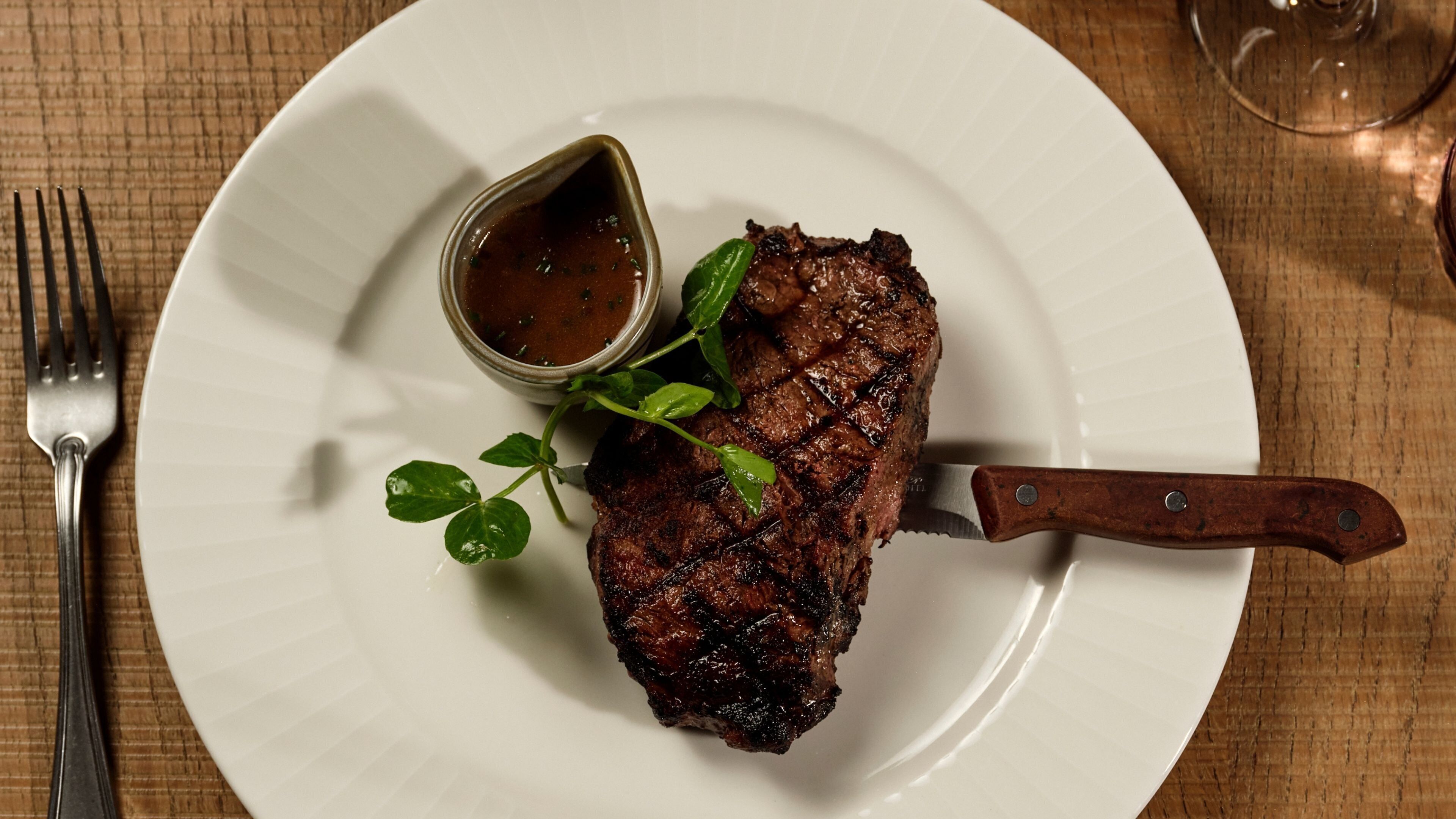 A steak with a knife on a plate, a fork, and a small bowl of sauce.