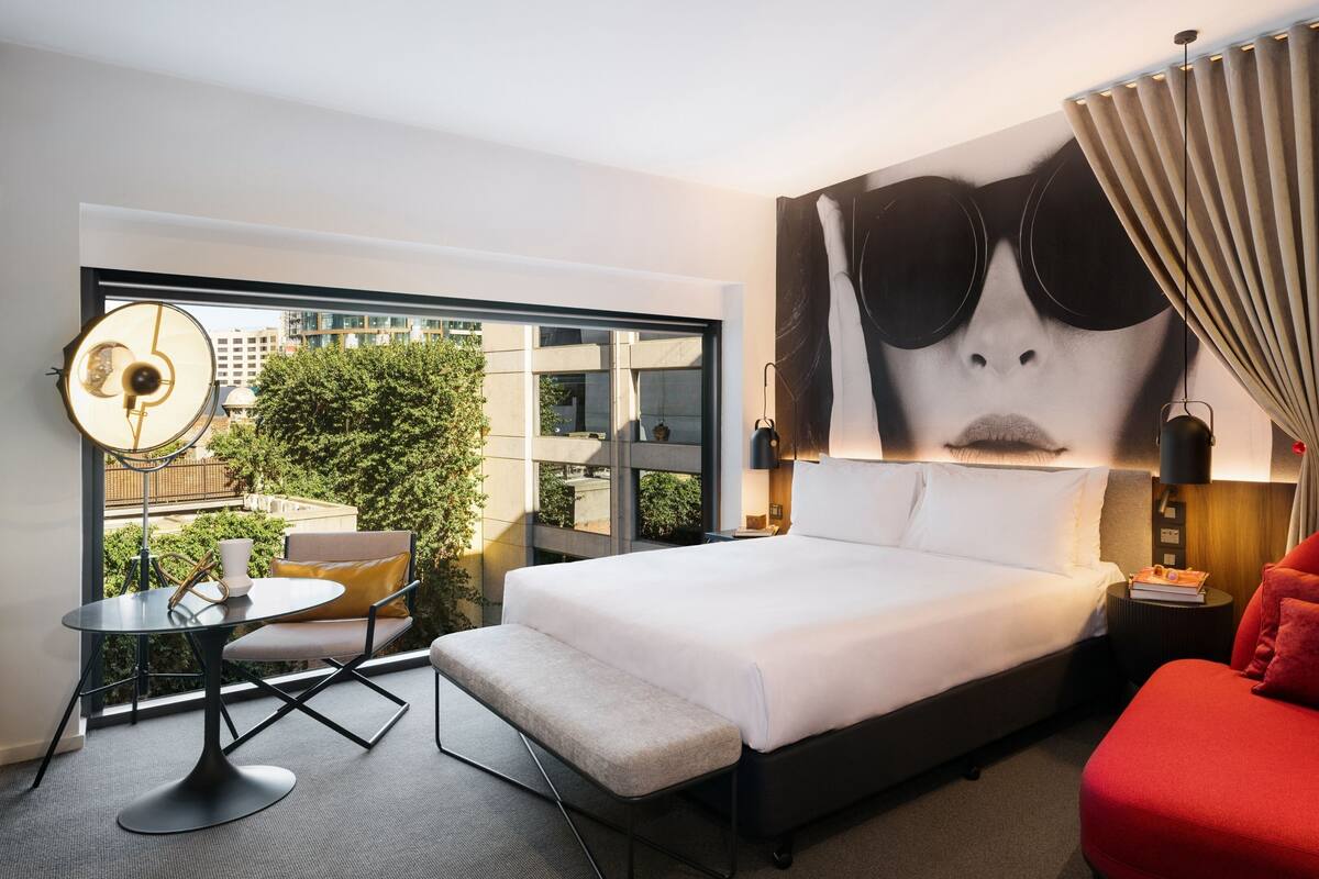 Ein Hotelzimmer mit einem großen Bett, einem roten Sessel, einem kleinen Tisch mit Stuhl, einem großen Fenster mit Blick auf Gebäude und Grünflächen sowie einer Wand mit einem großen abstrakten Gemälde.