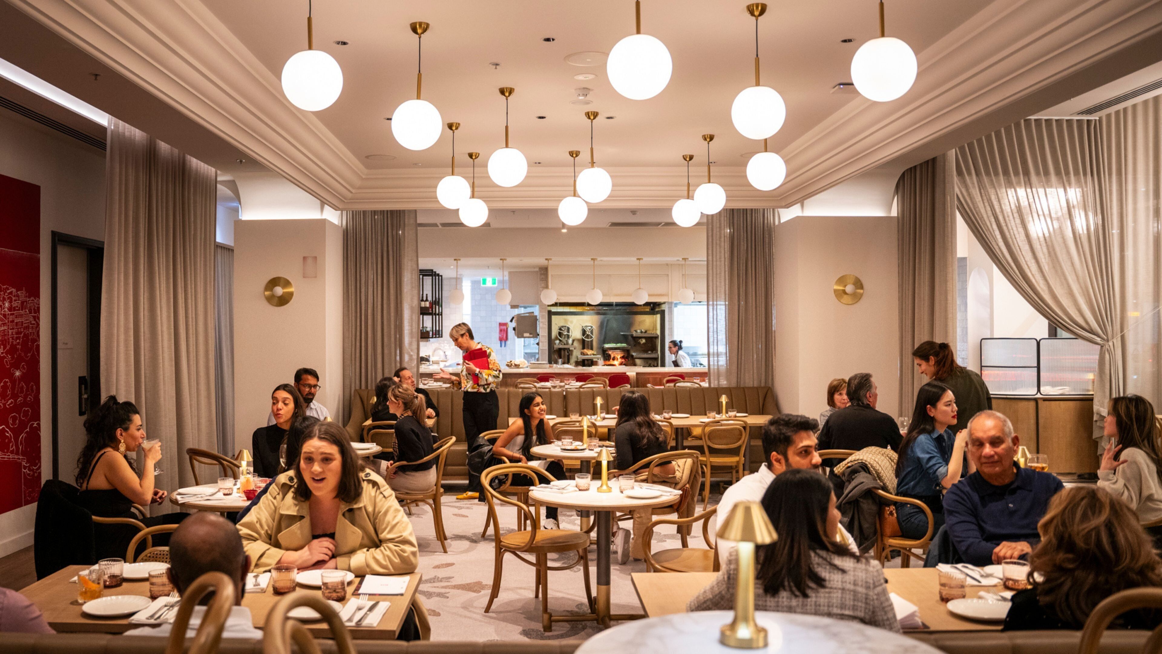 A spacious restaurant with round tables and chairs, a bar area in the background, and a modern ceiling design.