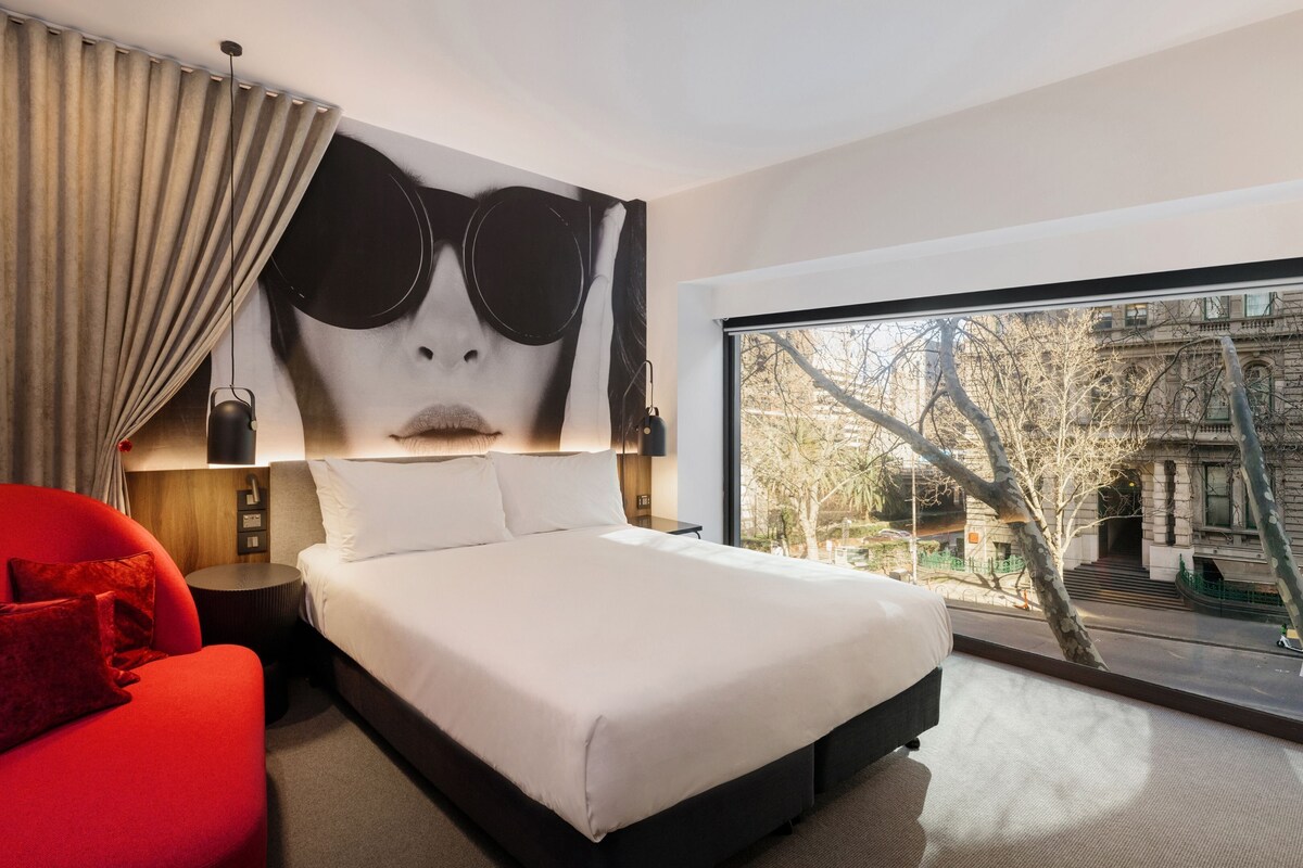 Una habitación de hotel moderna con una cama grande, un sillón rojo, vista a la ciudad a través de un ventanal amplio y una pieza de arte mural original.