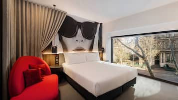 Una habitación de hotel moderna con una cama grande, un sillón rojo, vista a la ciudad a través de un ventanal amplio y una pieza de arte mural original.