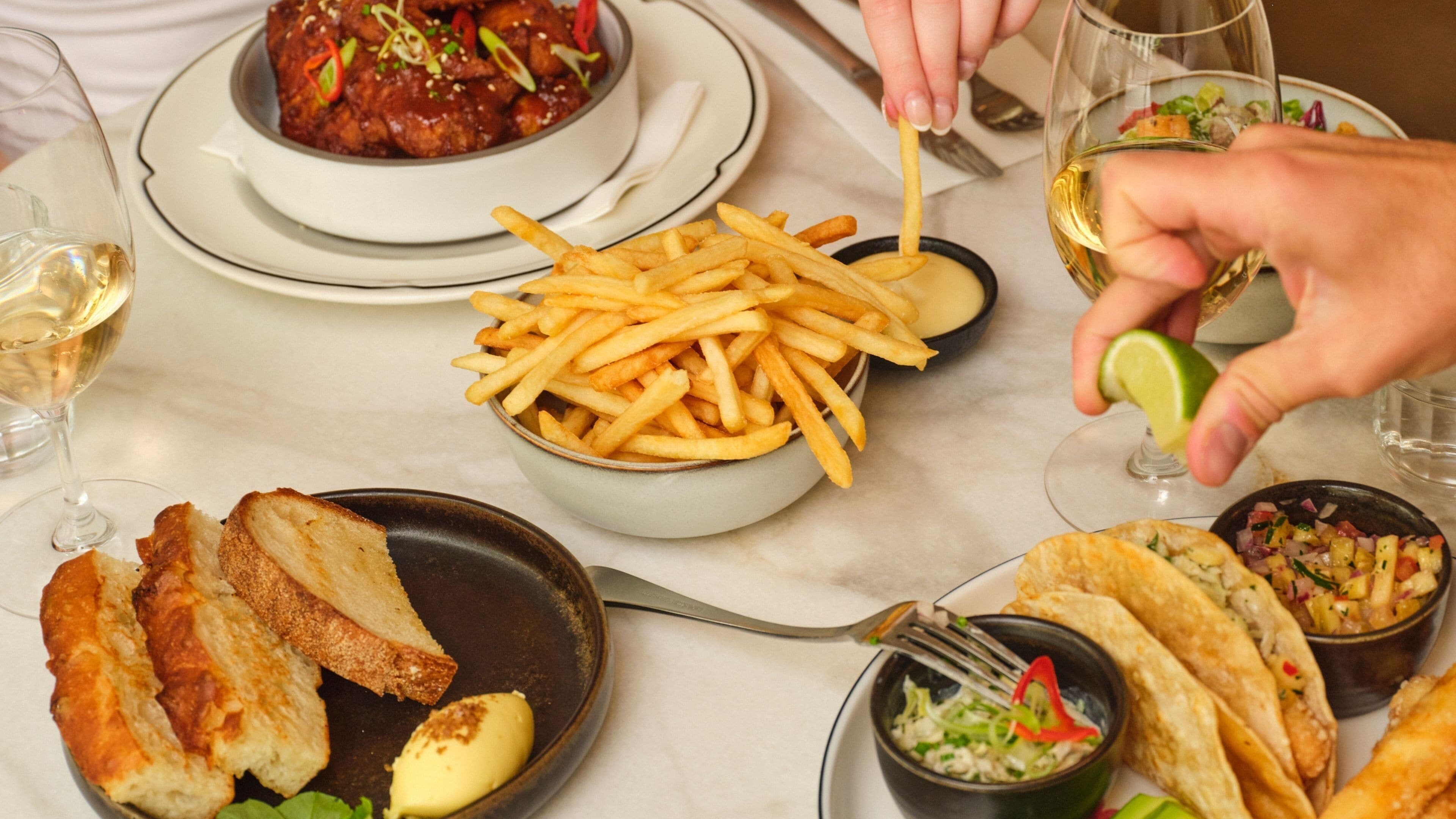 A dining table with various dishes including fries, bread, and tacos, along with glasses of wine.