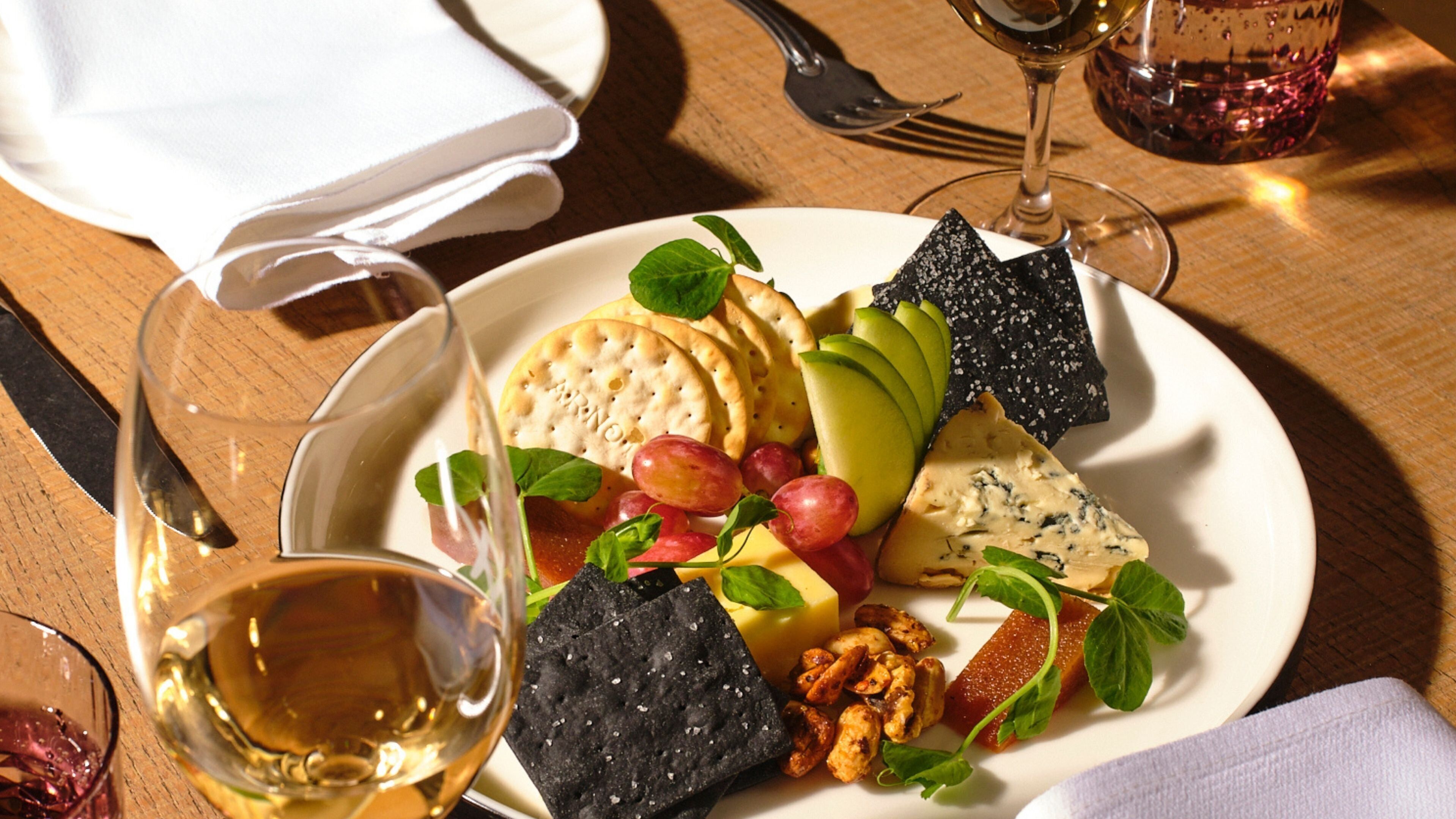 Un plato de queso, frutas y galletas sobre una mesa de madera, junto a una copa de vino blanco.