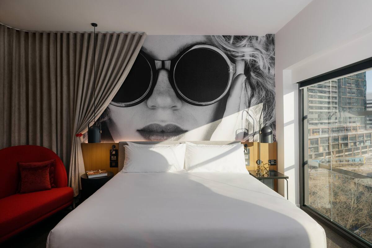 Ein Hotelzimmer mit einem großen Bett, einem roten Sessel, einem Nachttisch, einer Lampe und Blick auf eine Stadtlandschaft durch ein großes Fenster.