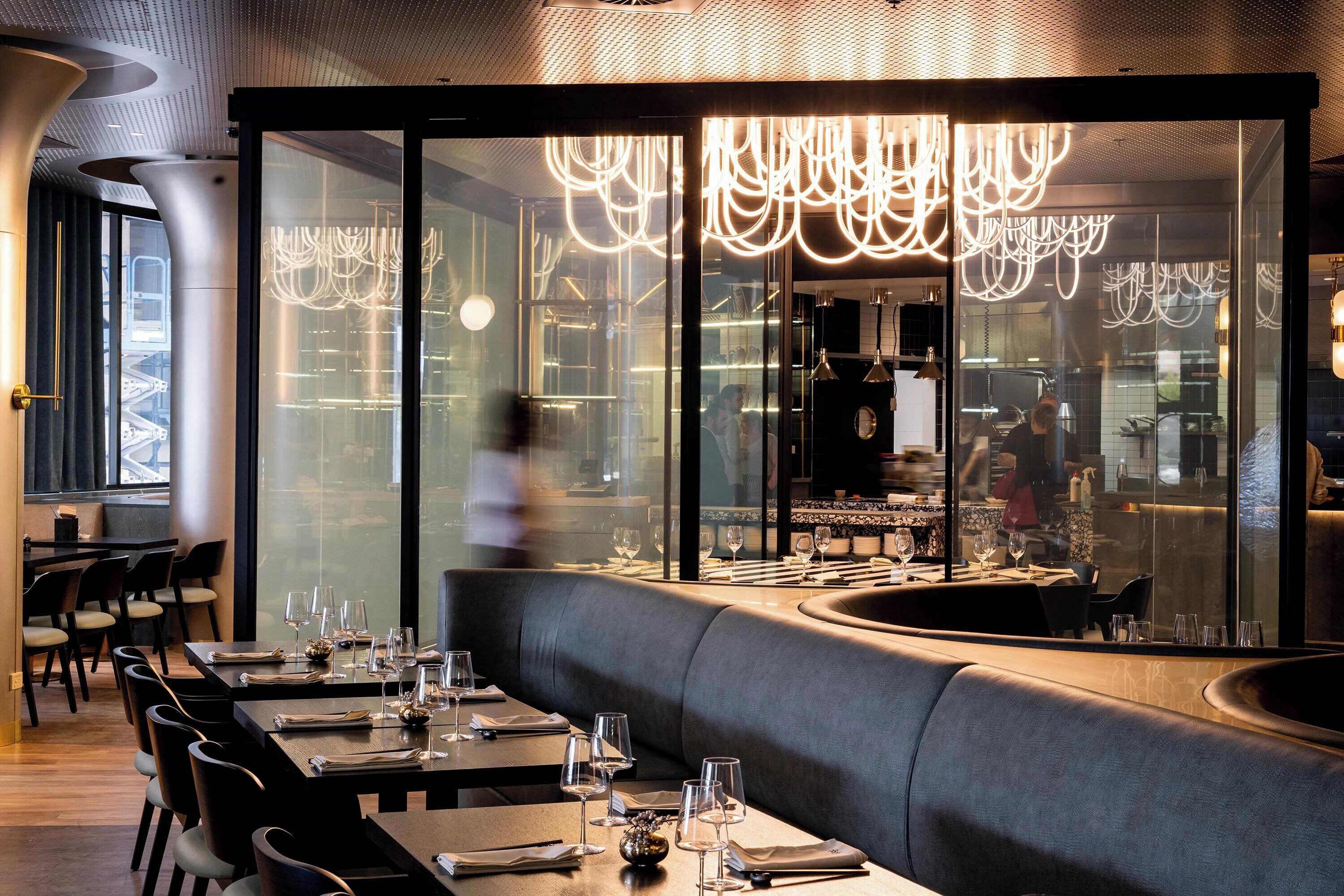 Un restaurant au décor moderne, avec un long comptoir agrémenté de tables et de chaises, un espace central avec un banc rembourré et une cloison en verre qui sépare la salle à manger de la cuisine.