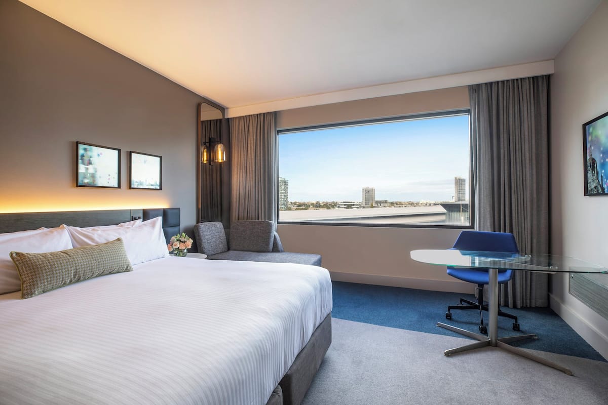 A hotel room with a large bed, a desk with a chair, a view of the city, and a window with curtains.
