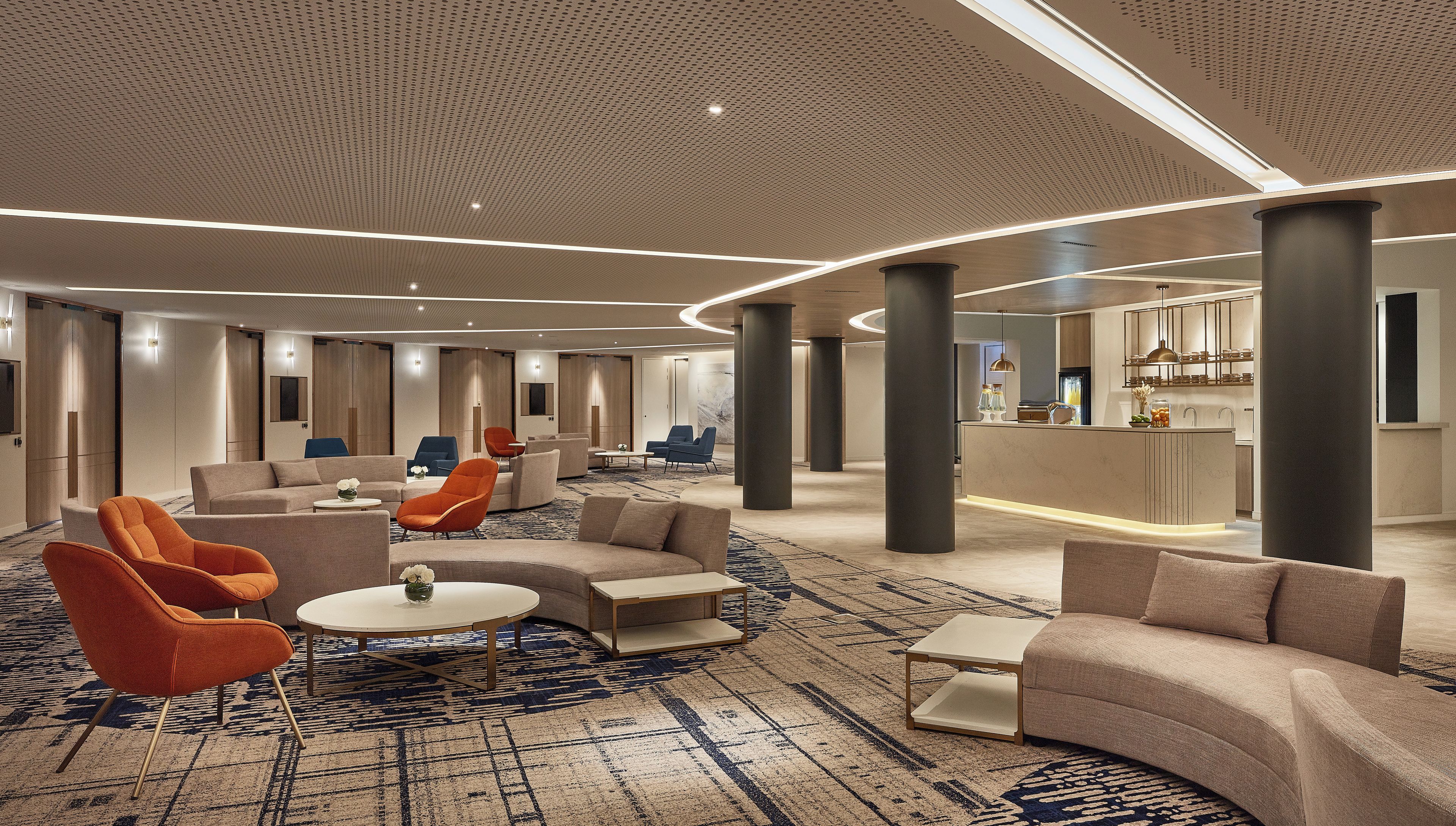 Un moderno vestíbulo de hotel con una zona de estar central, un bar y una cocina a la vista en el fondo.