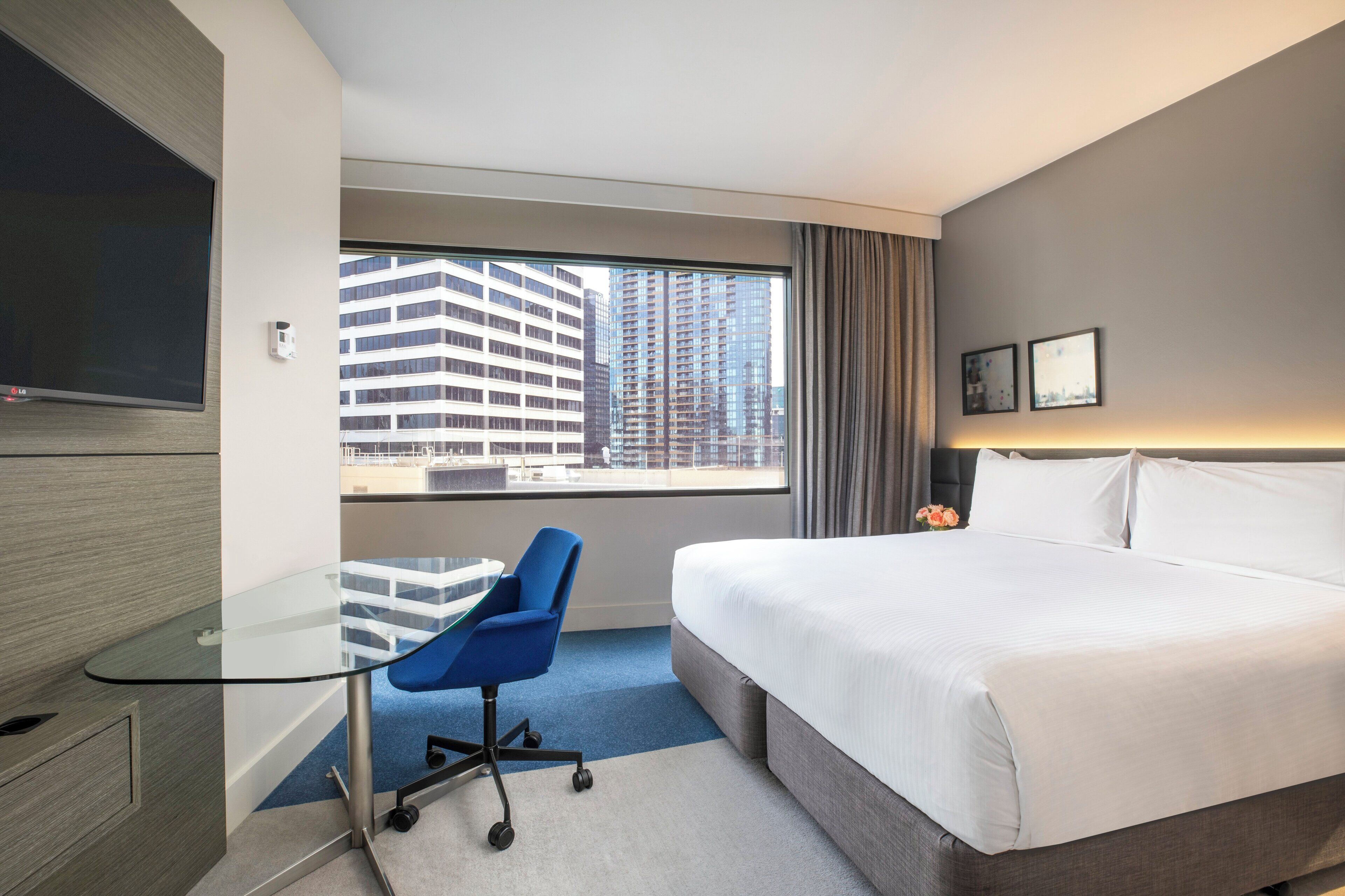 A hotel room with a large bed, a desk with a chair, a TV, and a window with a city view.