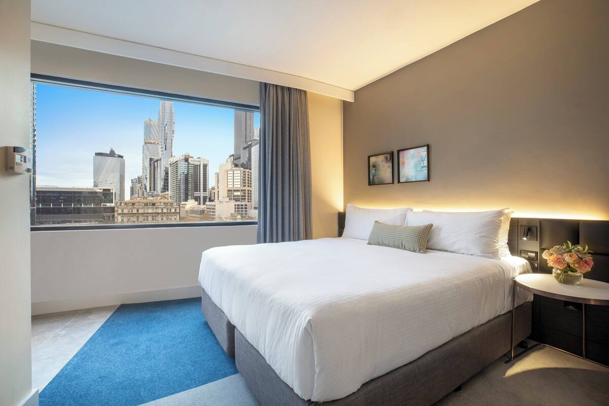 A hotel room with a large bed, a nightstand, a window with a city view, and a wall-mounted lamp.