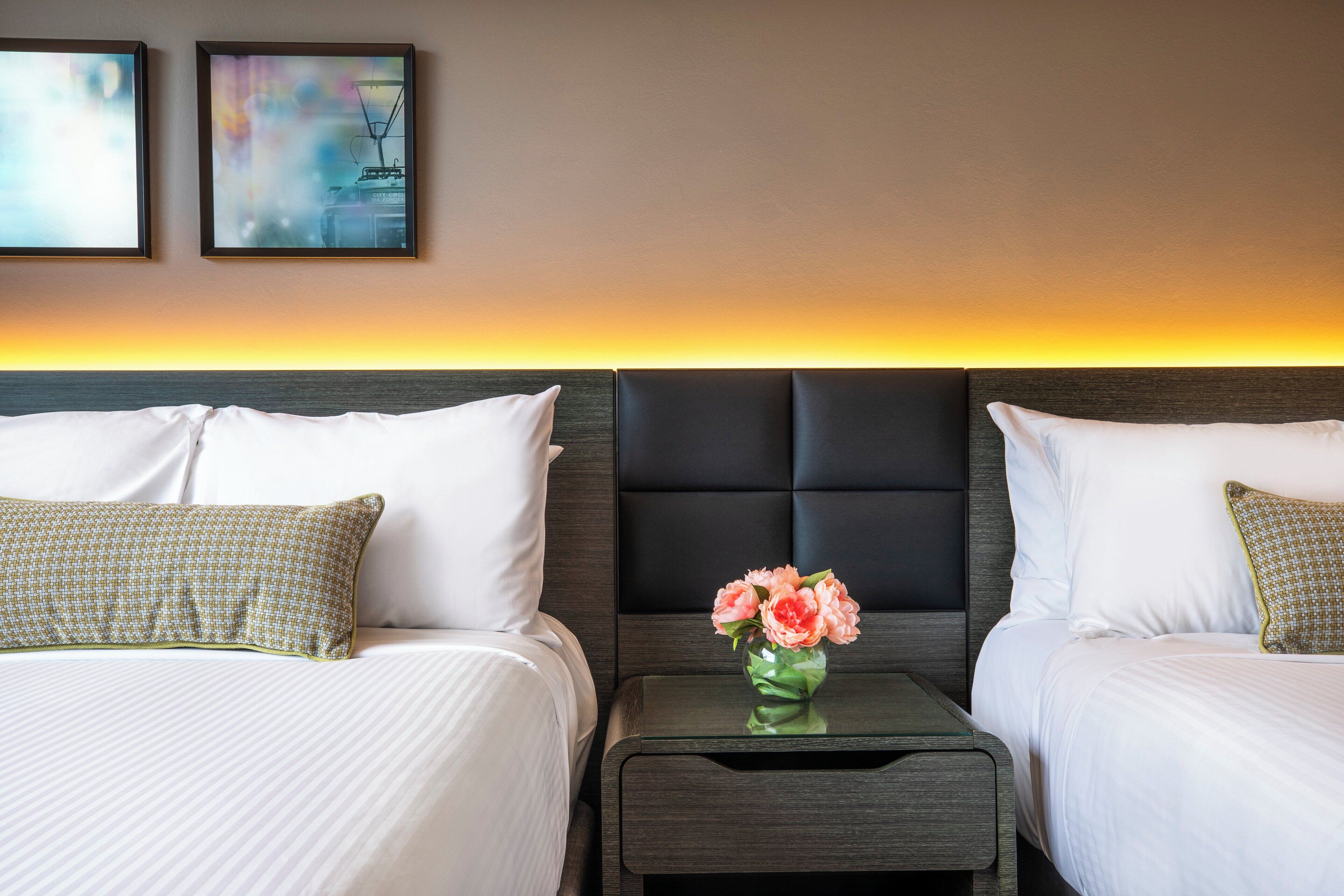 A hotel room with two beds, a nightstand with a vase of flowers, and two framed pictures on the wall.