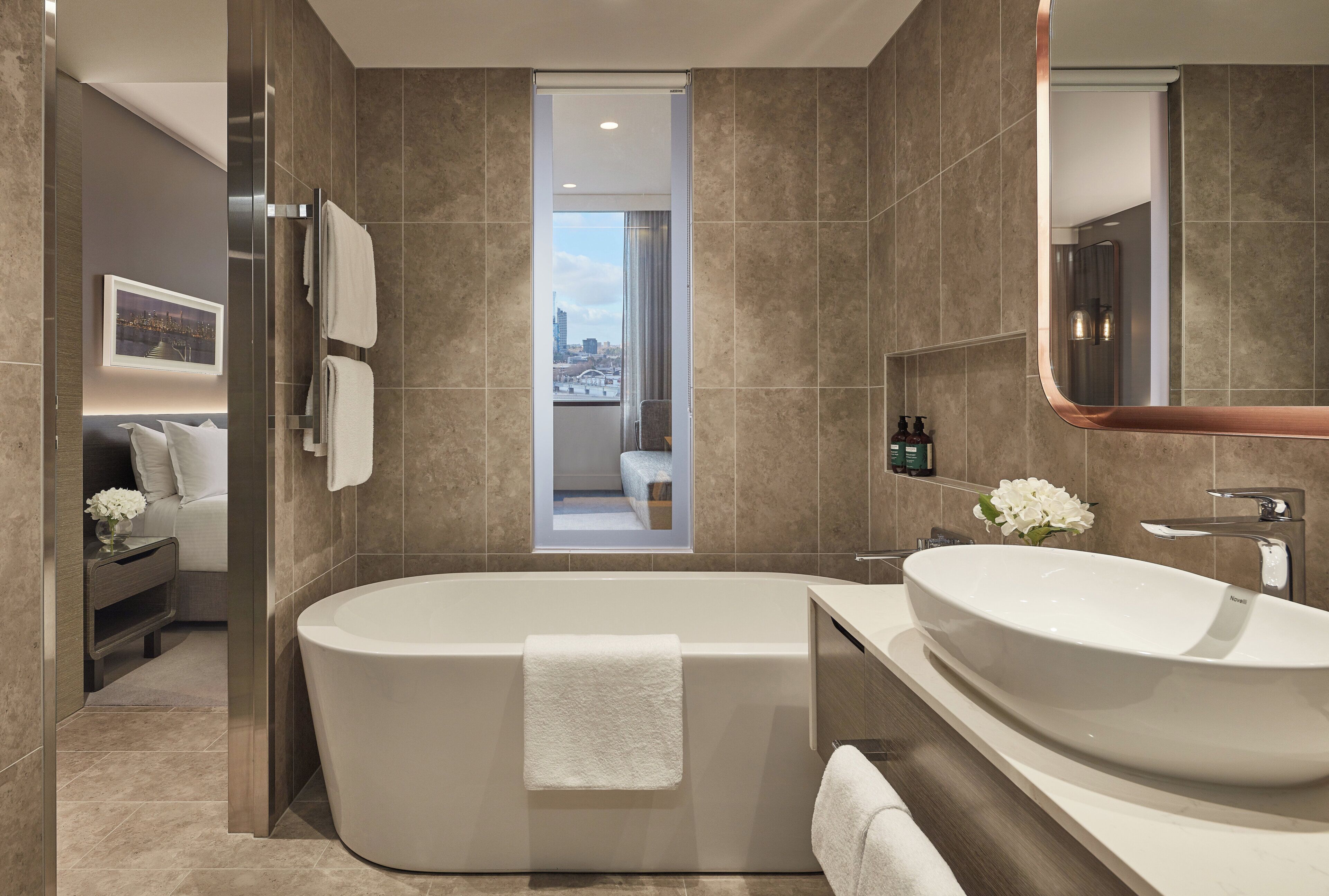 A modern bathroom with a freestanding bathtub, a large vanity with a mirror, and a view of a cityscape through a doorway.