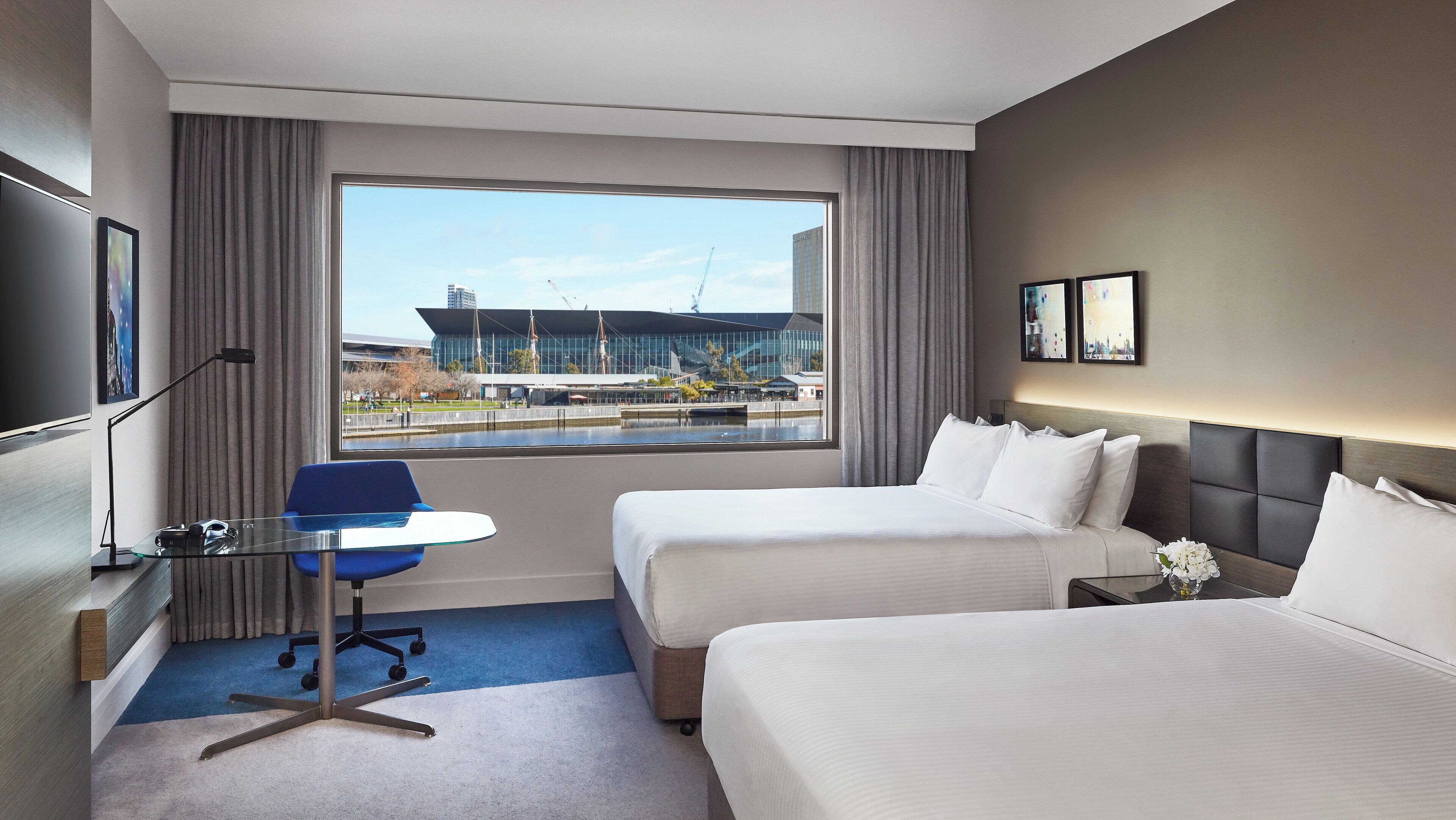 Habitación de hotel con dos camas, un escritorio, una silla, un televisor y un ventanal con vistas a un edificio moderno y una masa de agua.