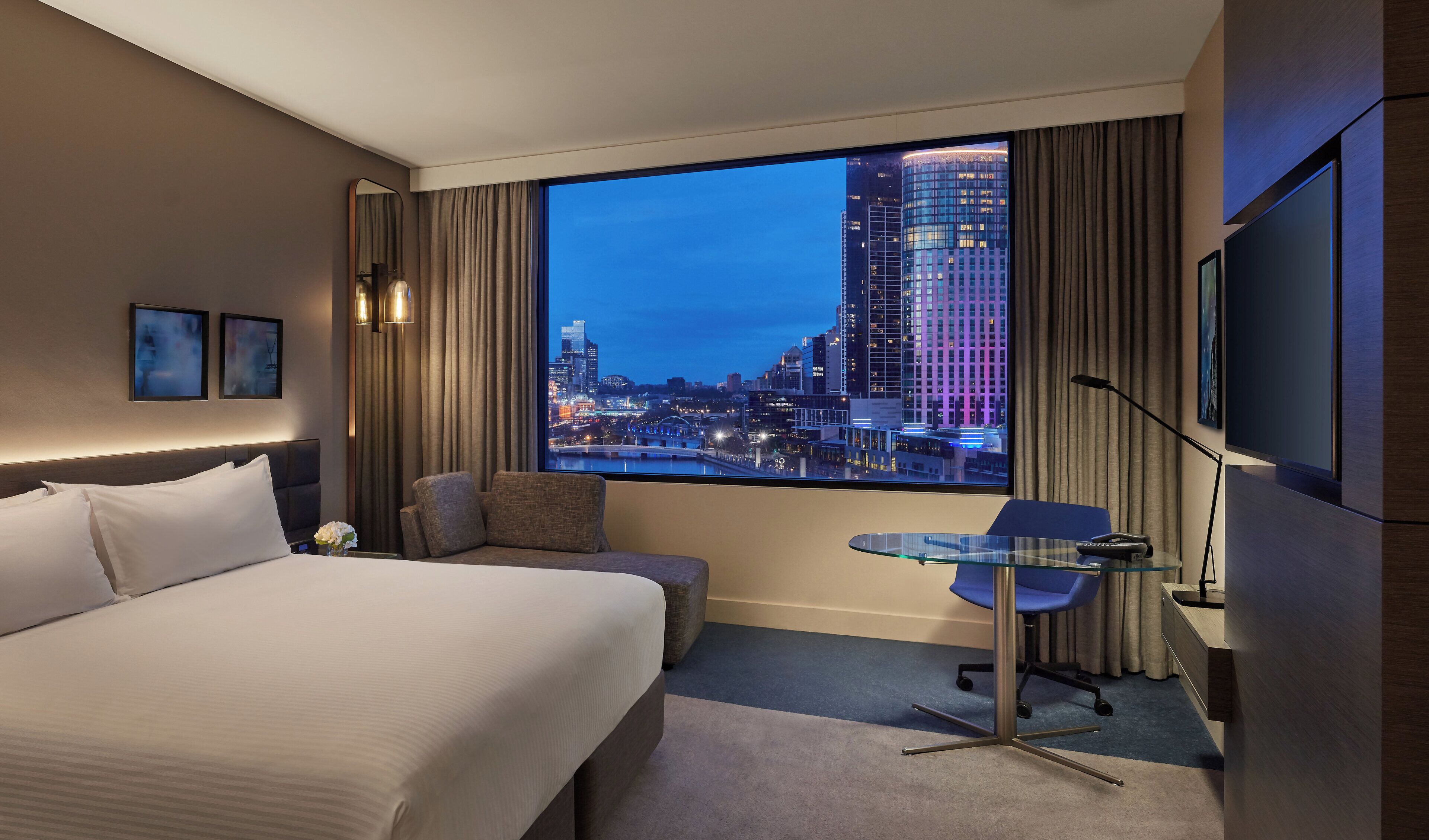 A hotel room with a large bed, a seating area, a small table, and a view of a cityscape at dusk.