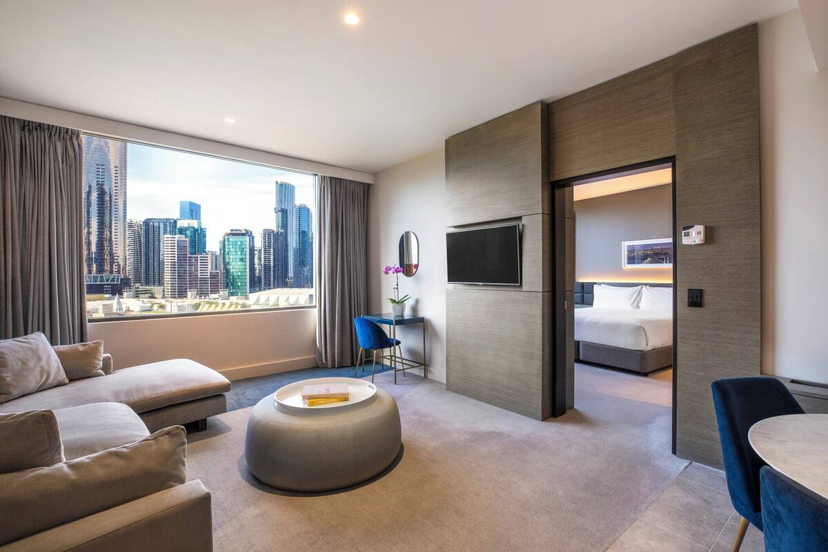 A modern hotel room with a large window offering a city view, a sofa, a round ottoman, a small desk, and a bed.