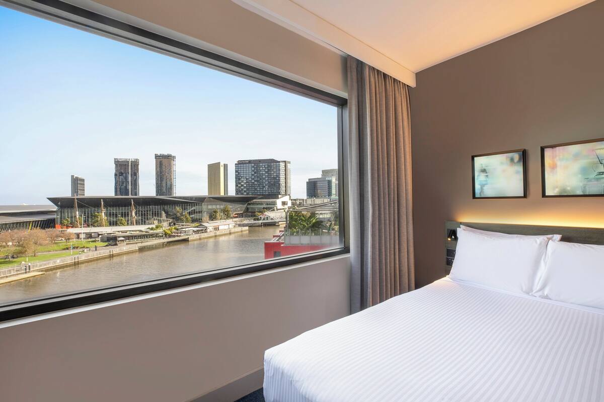 A hotel room with a large window offering a view of a city skyline and a river.