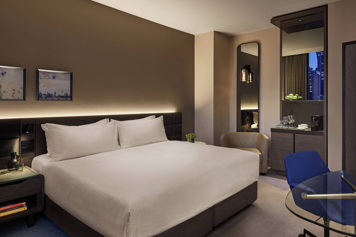 A modern hotel room with a large bed, bedside tables, a desk with a chair, a mirror, and artwork on the wall.