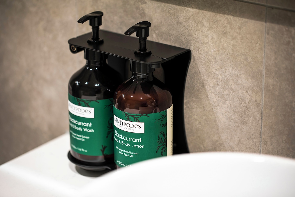Two bottles of Antipodes body wash and lotion on a black soap dispenser in a bathroom.