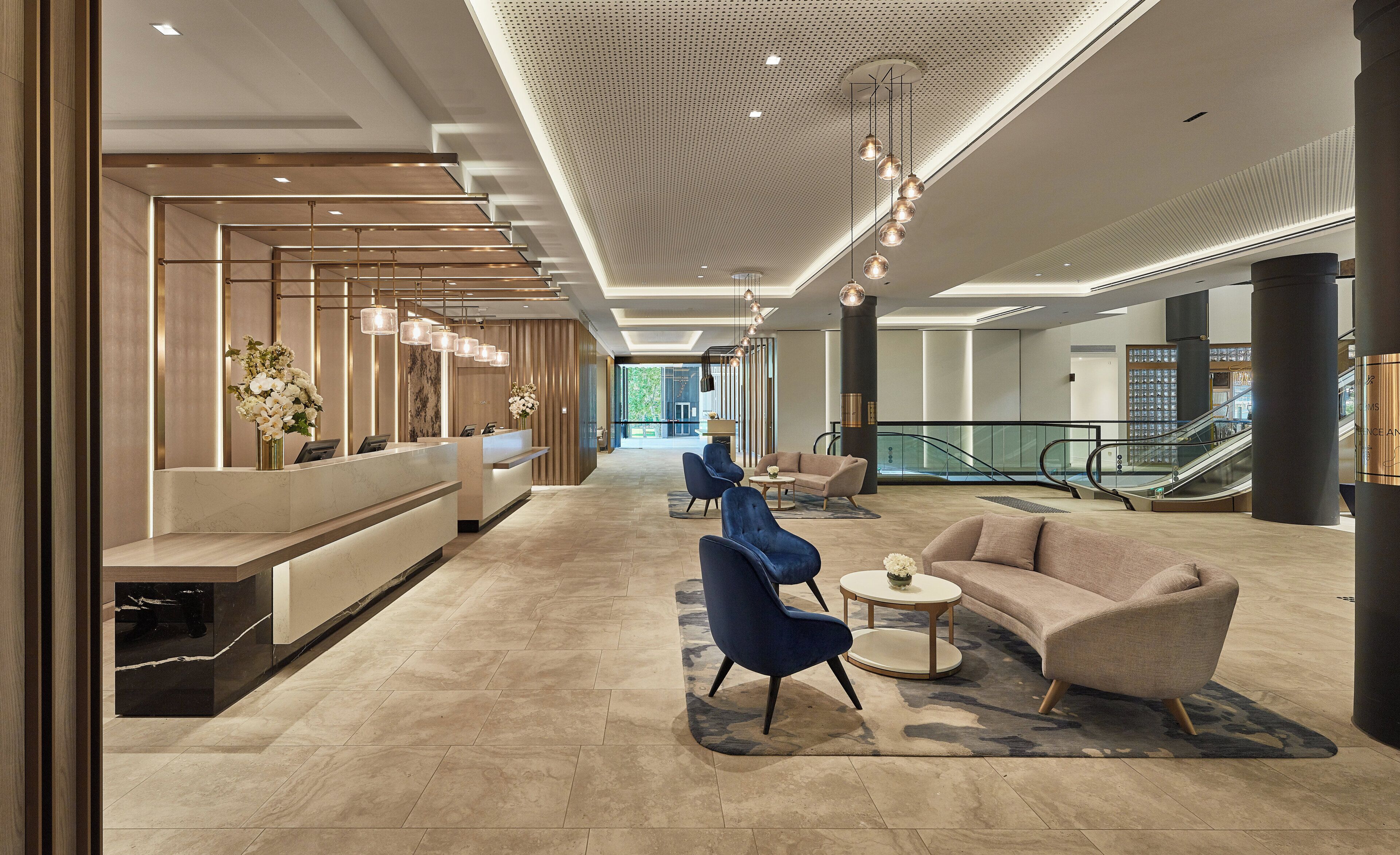 A modern hotel lobby with a reception desk, seating area, and an escalator.