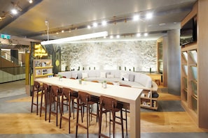 A modern dining area with a long table and wooden chairs, a white sofa, and a stone wall backdrop. - Crowne Plaza Melbourne by IHG (Docklands)