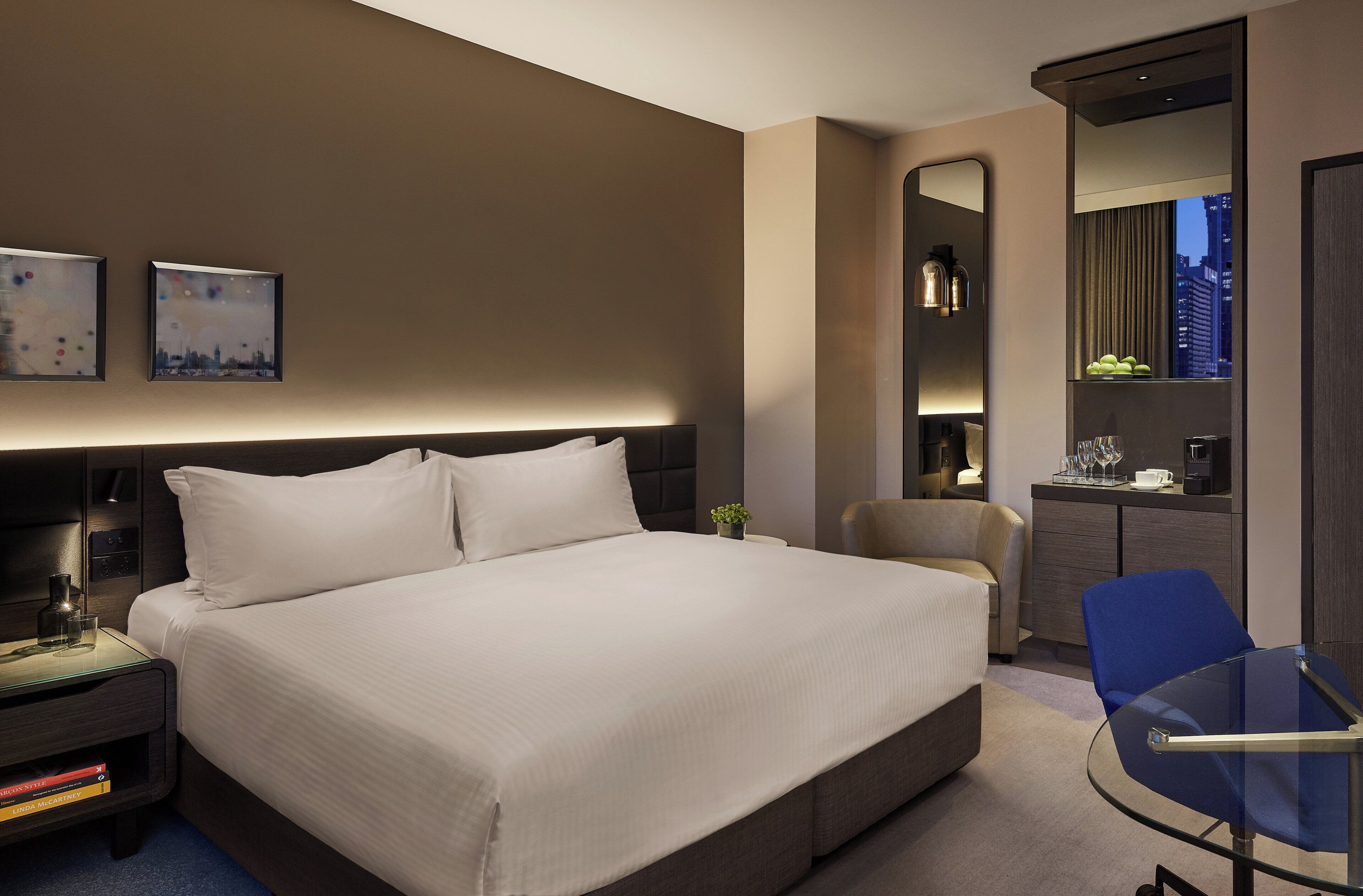 A modern hotel room with a large bed, bedside tables, a desk, a chair, and a mirror.
